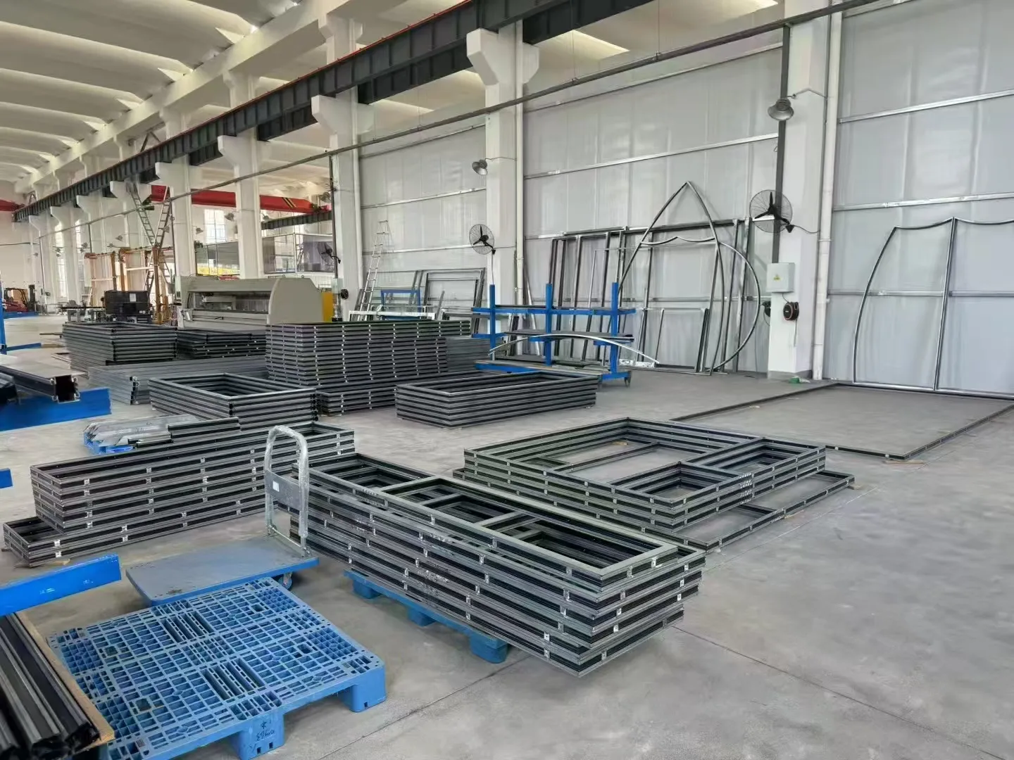 Steel window and door frames stacked in the workshop before glazing, with curved profiles for arched units leaning against the racking behind. Straightening, welding and surface treatment at this stage are what decide whether a frame is still true in fifty years.